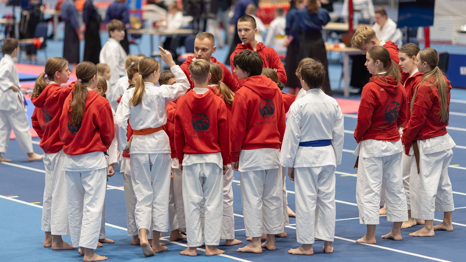 Trener Raion Fight Club w kręgu kilkunastu dzieci w kimonach na hali sportowej, tuż przed startem na zawodach karate